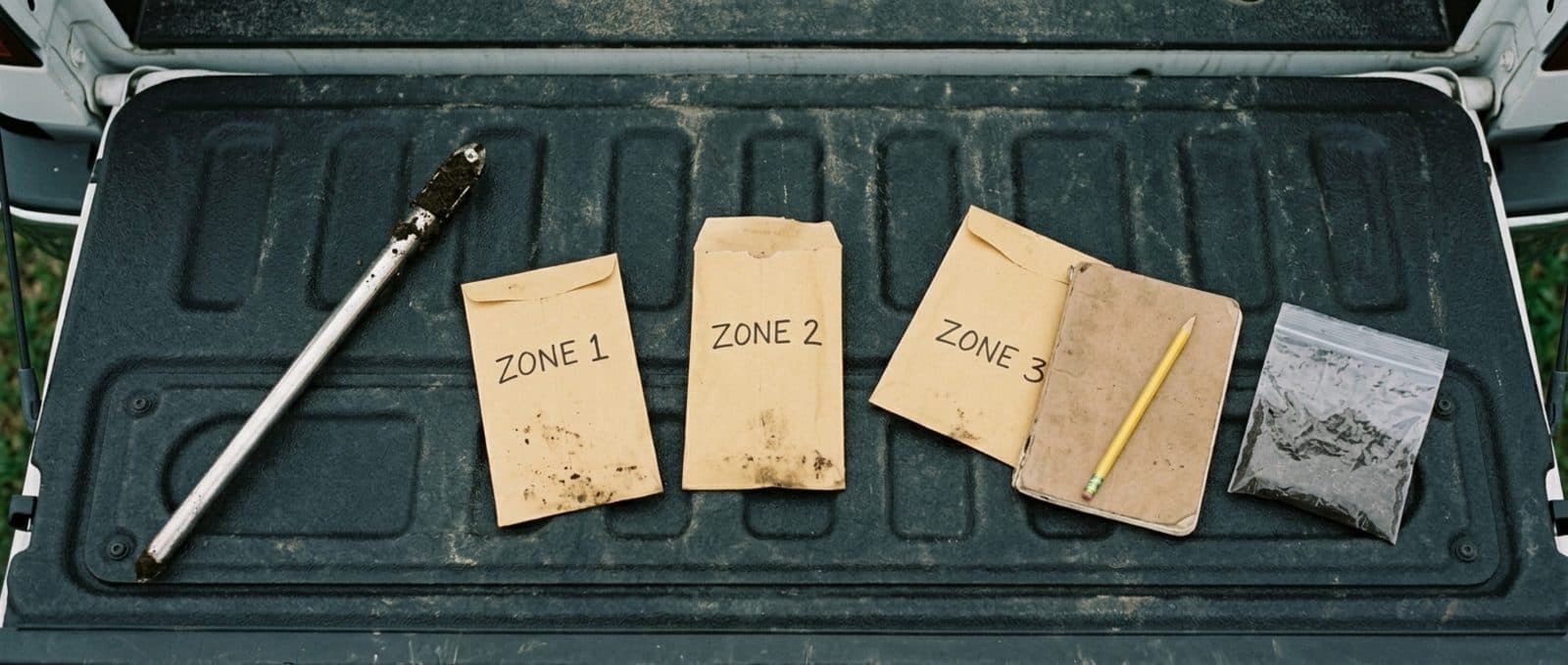 A weathered pickup tailgate photographed from overhead, with a stainless soil probe, three kraft-paper soil-sample envelopes labeled ZONE 1, ZONE 2, ZONE 3, a closed notebook with a yellow pencil, and a clear bag of dark loamy soil arranged in a horizontal line.